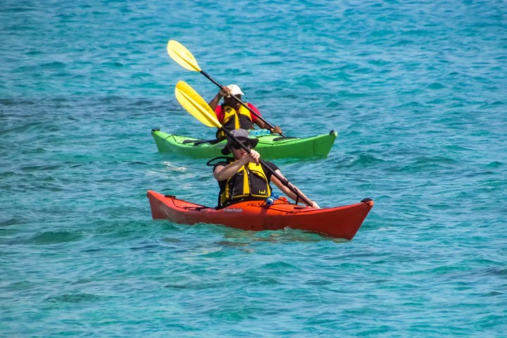 Two people kayaking on clear blue water, one in a red kayak and another in a green kayak.