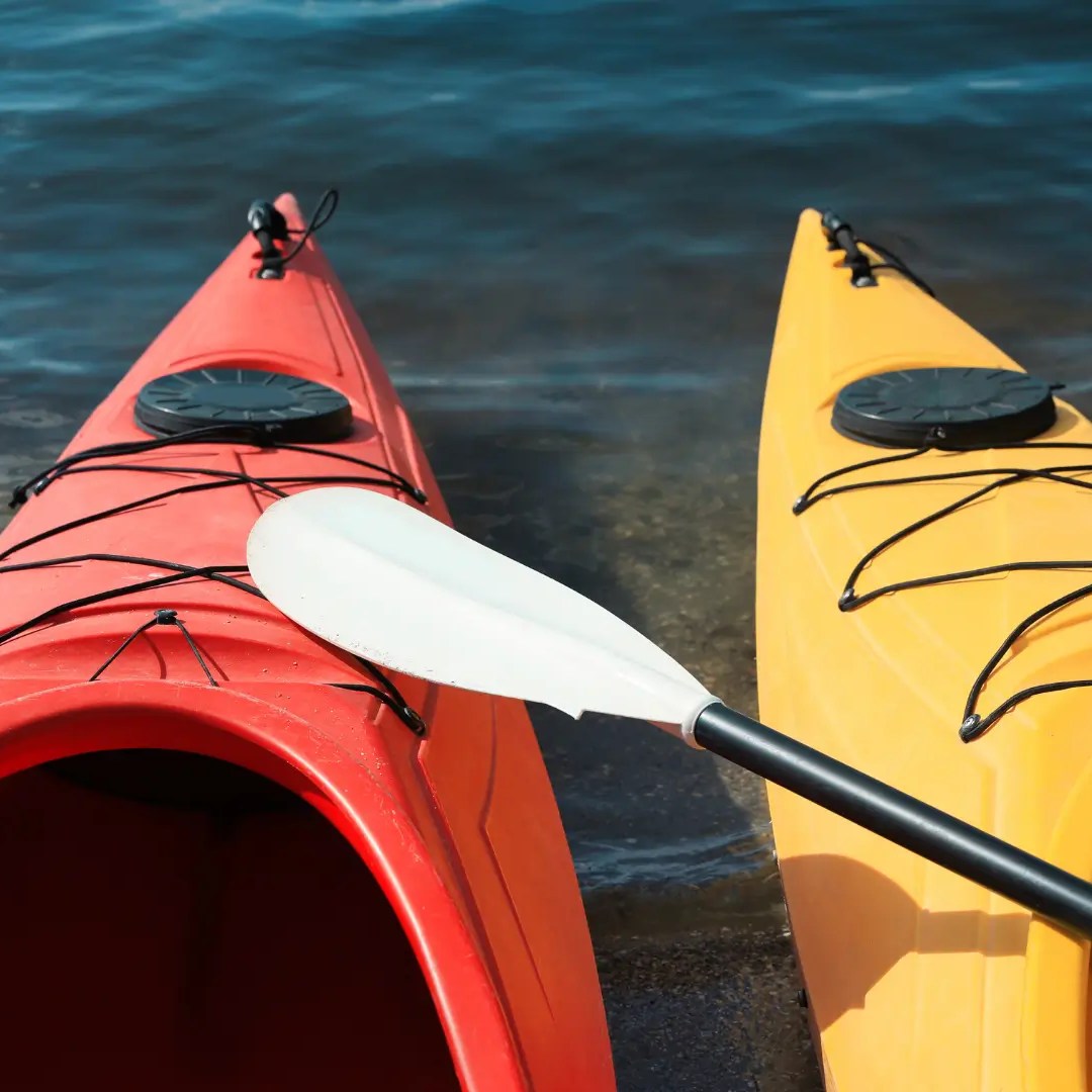 Two kayaks, one red and one yellow, on a shore with a paddle between them.