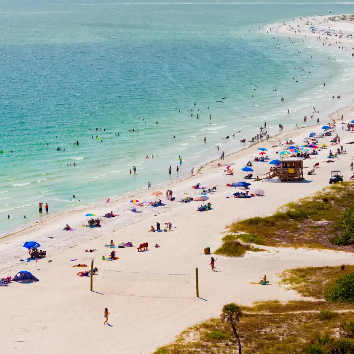 Busy beach with people, umbrellas, and volleyball nets along the shoreline.