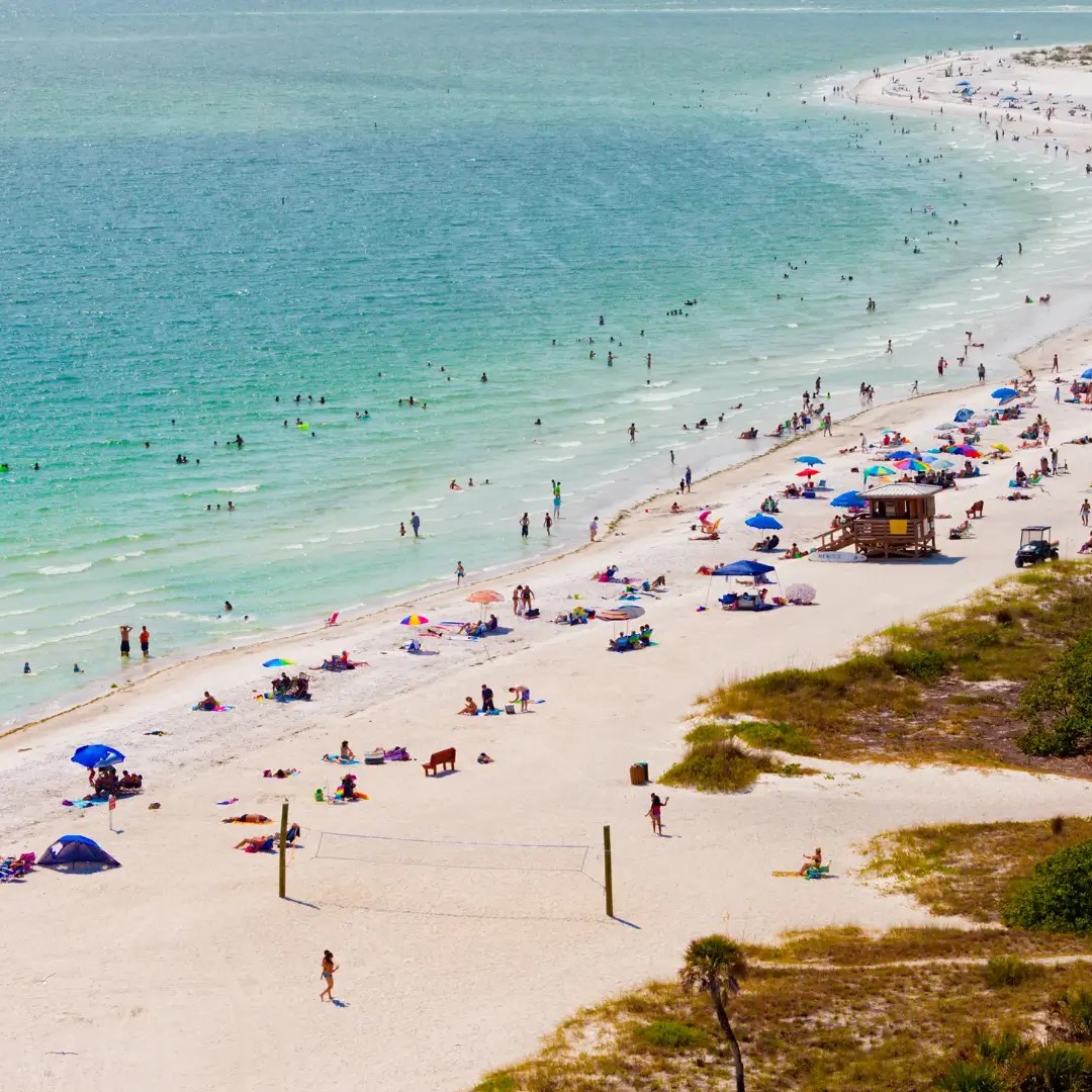 Busy beach with people, umbrellas, and volleyball nets along the shoreline.