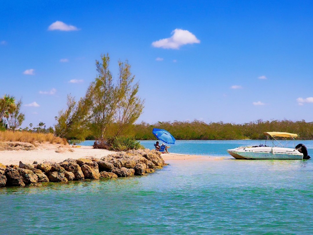 Beach scene with a boat and people under a blue umbrella on a sandy shore.