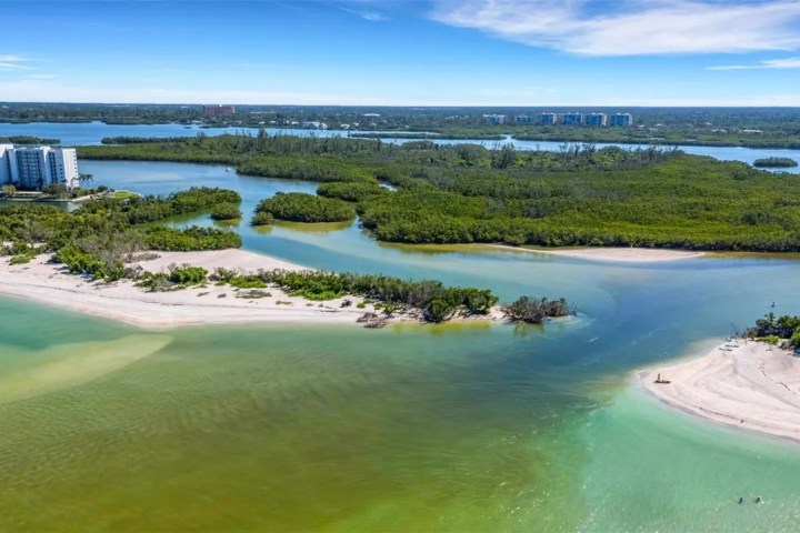 Aerial view of a beach, inlet, and dense greenery with buildings in the background.