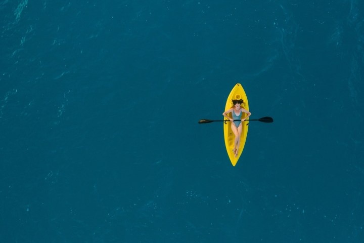 Woman in a yellow kayak on calm blue water, viewed from above.